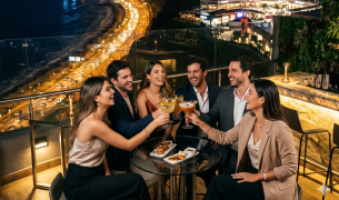 Grupo de amigos brindando con cócteles en un bar rooftop de Miraflores con vista nocturna a la Costa Verde iluminada y al centro comercial Larcomar.