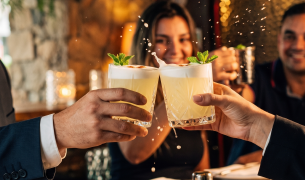 Primer plano de tres personas haciendo un brindis con vasos de Pisco Sour decorados con hojas de menta en un bar de Lima.